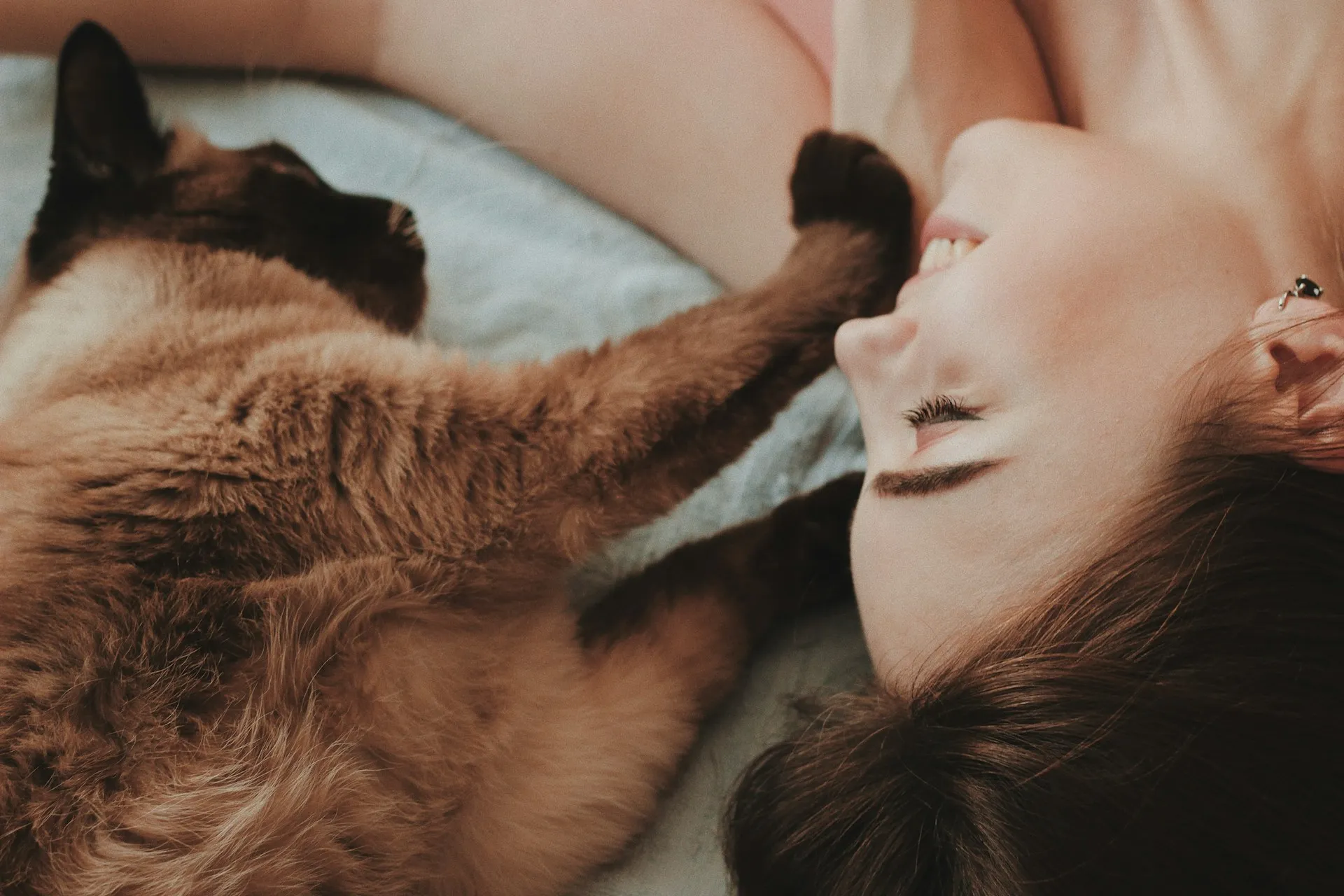 A close-up, overhead shot of a woman lying down and smiling at a Siamese cat, which has its dark paw gently resting near her face.